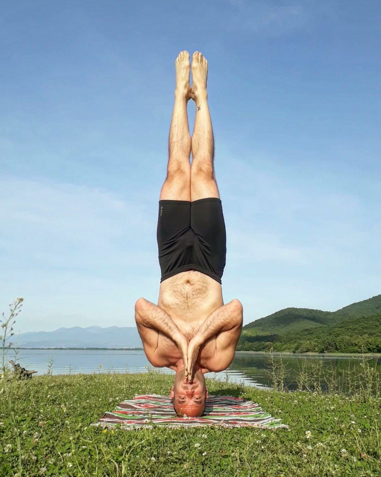 Man practicing yoga headstand pose on grass by serene Lake Kerkini with mountain views